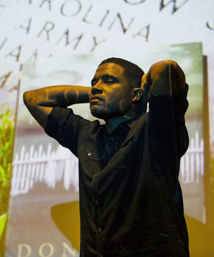 A photo of Mike Wiley on stage in BLOOD DONE SIGN MY NAME, hands clasped behind his head with stage projections across his face.