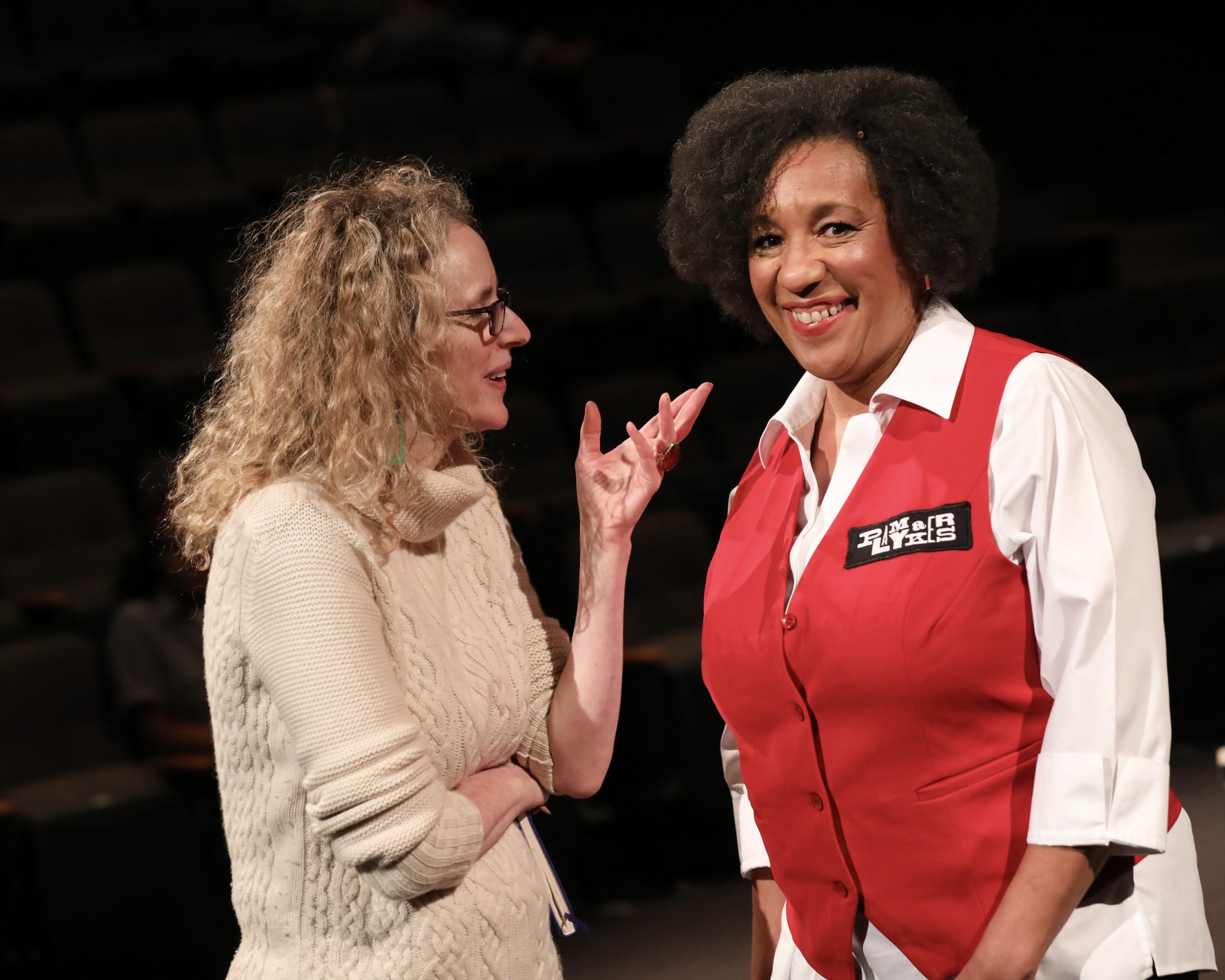 On the right, Kathy Williams, in costume as a PlayMakers usher, smiles directly at the camera. On the Left: Vivienne Benesch speaks with her.